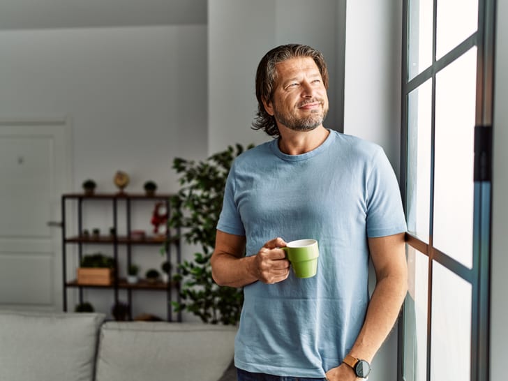 Mann steht in einem hellen Wohnraum am Fenster, hält eine Tasse in der Hand und blickt nach draussen; im Hintergrund sind Sofa, Regal und Pflanzen zu sehen.