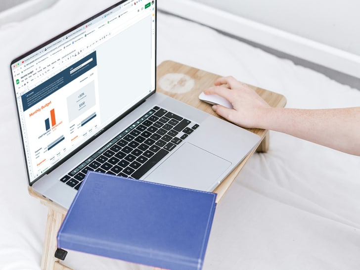 Person arbeitet an einem Laptop auf einem kleinen Tisch, daneben ein blaues Buch; auf dem Bildschirm ist eine Budgetübersicht mit Diagrammen zu sehen.
